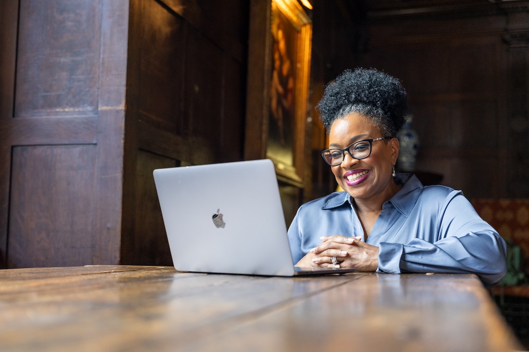 Woman seated, smiling, looking at her laptop.